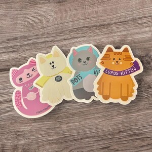Pu&ograve; includere: Quattro adesivi di gatto colorati con messaggi diversi. Il gatto rosa dice "Thrifty", il gatto bianco dice "CUDDLY PUPPY", il gatto grigio dice "POTS" e il gatto arancione dice "LUPUS KITTY!"