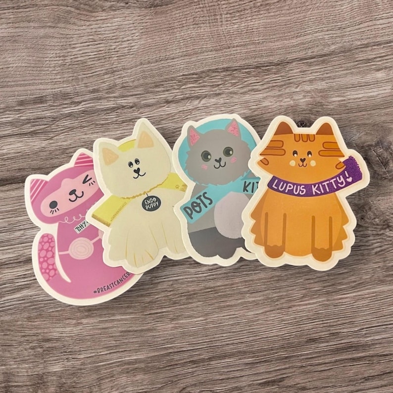 Pu&ograve; includere: Quattro adesivi di gatto colorati con messaggi diversi. Il gatto rosa dice "Thrifty", il gatto bianco dice "CUDDLY PUPPY", il gatto grigio dice "POTS" e il gatto arancione dice "LUPUS KITTY!"