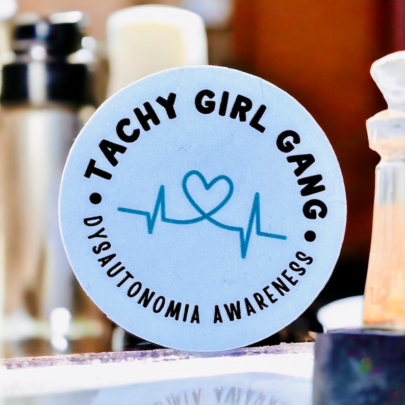 May include: A white round sticker with black text that reads "Tachy Girl Gang" and "Dysautonomia Awareness" around a blue heart and heartbeat graphic.