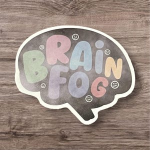 Könnte beinhalten: Ein Aufkleber in Form eines Gehirns mit den Worten "Brain Fog" in bunten, blasenförmigen Buchstaben. Der Aufkleber hat einen weißen Rand und befindet sich auf einem Holzgrund.