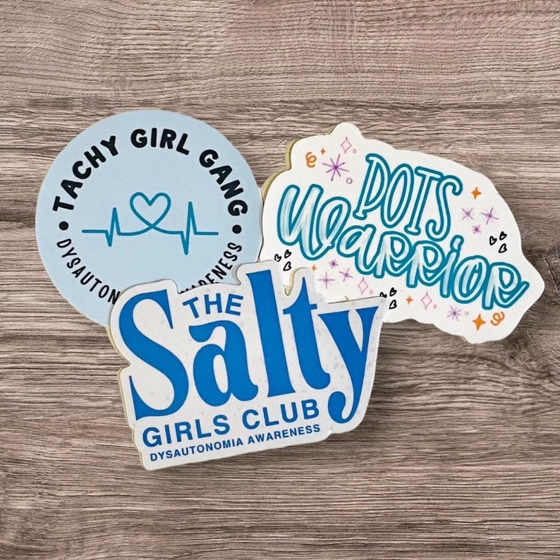 May include: Three stickers with blue text on a light wood background. The first sticker is light blue with a heart and a heartbeat line and the text "Tachy Girl Gang Dysautonomia Awareness". The second sticker is white with blue text and the words "The Salty Girls Club Dysautonomia Awareness". The third sticker is white with blue text and the words "POTS Warrior" with pink and purple stars and hearts.