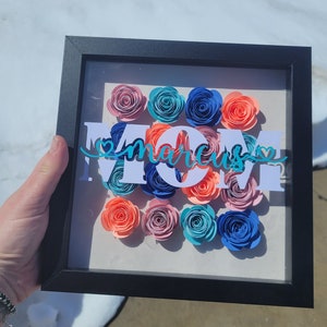 May include: A framed paper craft featuring the word "MOM" in white lettering and the name "marcus" in teal lettering. The frame is black and holds an assortment of paper flowers in shades of pink, blue, and orange.