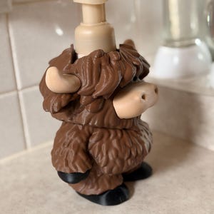May include: A novelty soap dispenser in the shape of a Highland cow. The dispenser is brown with a beige pump and features a detailed design of the cow's fur, horns, and nose. The cow has black hooves. This dispenser adds a whimsical touch to any bathroom.