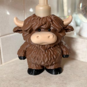 May include: A novelty soap dispenser in the shape of a Highland cow. The dispenser is brown with a tan pump, face, horns, and hooves. The cow's fur is a darker shade of brown. It stands on a white surface.