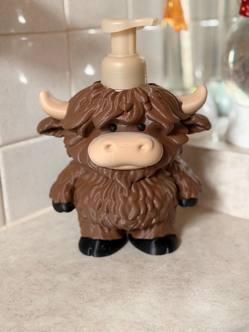 May include: A novelty soap dispenser in the shape of a Highland cow. The dispenser is brown with a tan pump, face, horns, and hooves. The cow's fur is a darker shade of brown. It stands on a white surface.