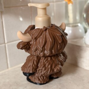 May include: A brown soap dispenser in the shape of a Highland cow. It features a beige pump and horns, with a textured, shaggy coat. The base is black. The dispenser is designed for a countertop.