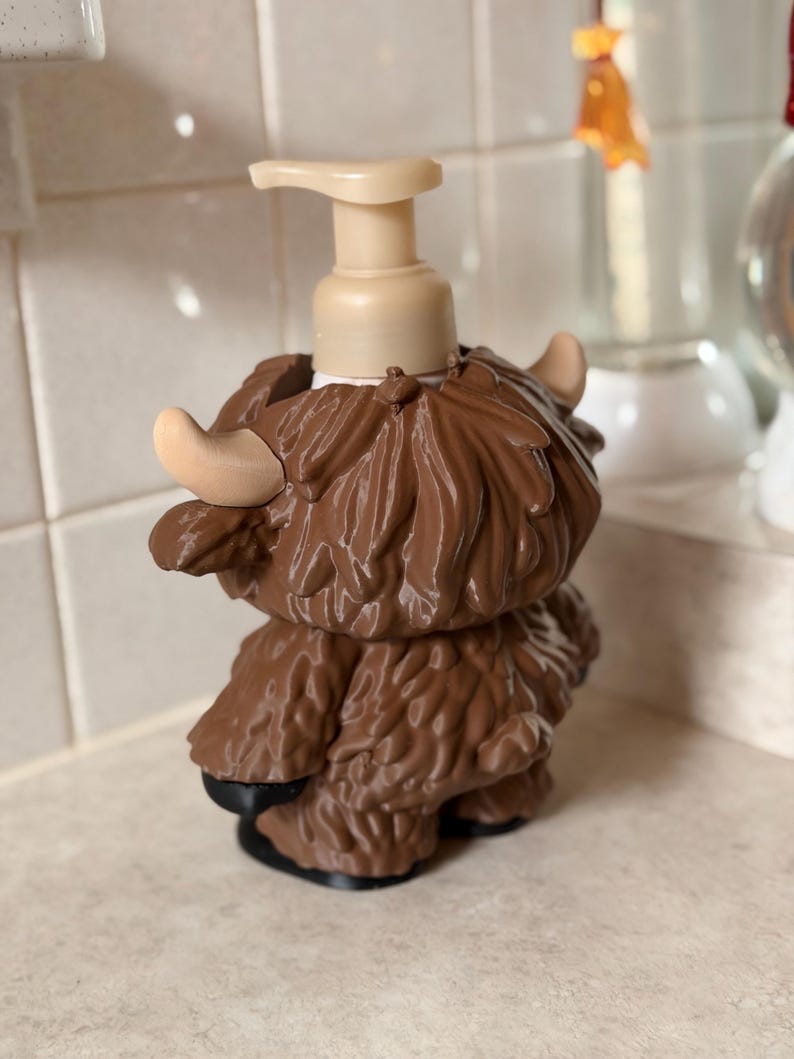 May include: A brown soap dispenser in the shape of a Highland cow. It features a beige pump and horns, with a textured, shaggy coat. The base is black. The dispenser is designed for a countertop.