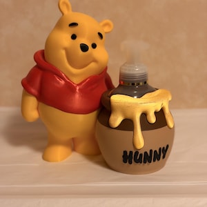 May include: A yellow Winnie the Pooh figurine wearing a red shirt stands next to a brown honey pot-shaped soap dispenser. The dispenser has a gold-colored honey pour and the word "HUNNY" in black.