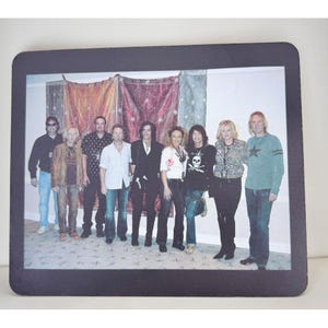 May include: A rectangular mousepad featuring a group photograph of nine people standing in a line. The individuals are dressed in casual clothing, including jeans, shirts, and jackets, positioned in front of three decorative fabric wall hangings.