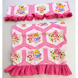 May include: A set of two fabric window valances featuring a repeating hexagonal pattern with pink lace-style borders. The print displays cartoon characters with strawberry motifs on a white background, finished with a solid dark pink ruffled hem.