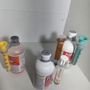 May include: Several bottles of children's medicine and syringes are arranged on a white surface. The bottles have white and orange labels. Syringes are yellow, brown, and teal. The medicine bottles are labeled with the brand name "Tylenol".