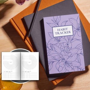May include: A purple habit tracker journal with a floral design. The cover features a stylized drawing of lilies in blue ink. The text "Habit Tracker" is printed in blue on a white rectangle.