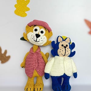 Puede incluir: Dos muñecos de ganchillo hechos a mano. Un muñeco es amarillo con un sombrero y chaleco rosa. El otro muñeco es azul con una chaqueta blanca. Ambos muñecos tienen rasgos faciales en blanco y negro y están de pie.