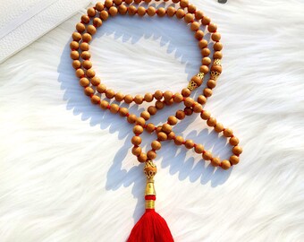 May include: A brown wooden mala bead necklace with a red tassel. The necklace is made of 108 beads and has a gold-colored metal charm at the end.
