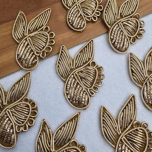 Op de afbeelding: Een verzameling goudkleurige geborduurde patches, elk met een gestileerd blad- of bloembladontwerp met ingewikkelde metalen draaddetails, gerangschikt in een verspreid patroon over een oppervlak met een witte stof en houttextuur.