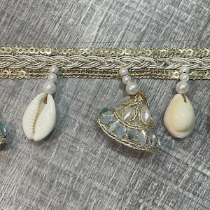 May include: Gold-colored trim with a woven design and attached embellishments. The trim features white seashells, pearl beads, and a decorative gold and silver pendant. The trim is on a gray wooden surface.