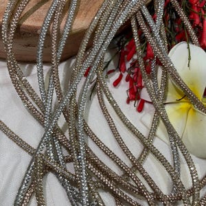 Può includere: Primo piano di più fili di finiture scintillanti con strass argento e oro. La finitura è disposta a strati, con uno sfondo bianco e fiori rossi sullo sfondo.