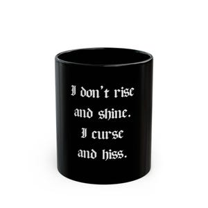 May include: Black ceramic mug with the text "I don't rise and shine. I curse and hiss." in white gothic font. The mug is cylindrical and has a glossy finish. A humorous and sarcastic gift.