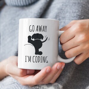 Peut inclure: Une tasse en céramique blanche avec une silhouette noire d'un chien tourné dos et la queue qui remue. Le texte "Go Away I'm Coding" est imprimé sur la tasse.