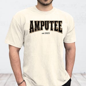 May include: Cream-colored t-shirt featuring the word "AMPUTEE" in bold, black letters with a leopard print fill. Below the word is "est 2025" in a smaller font. The shirt has short sleeves and a crew neck.