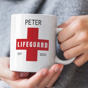 May include: White ceramic mug with the name "PETER" printed in black above a red cross design with the word "LIFEGUARD" in white. Below the cross are the words "EST" and "2023" in black. The mug has a white handle.