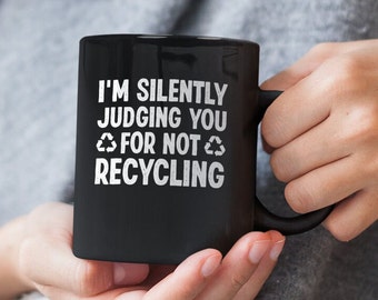 May include: Black coffee mug with white text that reads "I'm silently judging you for not recycling" with a recycle symbol on either side of the word "not".