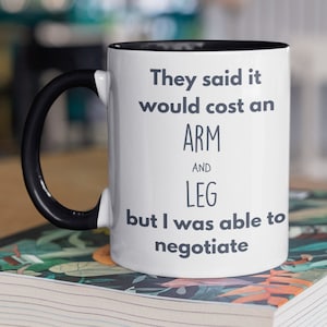 May include: White ceramic coffee mug with a black rim and handle. The mug has black text that reads "They said it would cost an ARM AND LEG but I was able to negotiate."