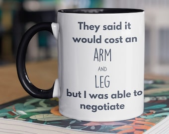 May include: White ceramic coffee mug with a black rim and handle. The mug has black text that reads "They said it would cost an ARM AND LEG but I was able to negotiate."