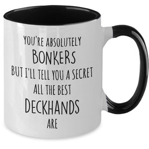 May include: White ceramic coffee mug with a black interior and handle. The mug has black text that reads "You're absolutely bonkers but I'll tell you a secret all the best deckhands are."