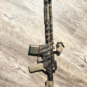 May include: A rifle with a camouflage pattern in shades of brown and black. It has a tan grip, stock, and various attachments, including a scope and a flashlight. The rifle is positioned diagonally on a wooden surface.