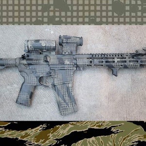 May include: A tactical rifle with a camouflage pattern in shades of grey, black, and tan. The rifle is equipped with a scope, a flashlight, and a vertical foregrip. The background is a concrete surface.