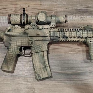 May include: A camouflaged rifle with a scope, flashlight, and vertical grip. The rifle has a magazine and is painted in shades of green, brown, and tan. The stock and handguard are also camouflaged. The rifle is lying on a wooden surface.