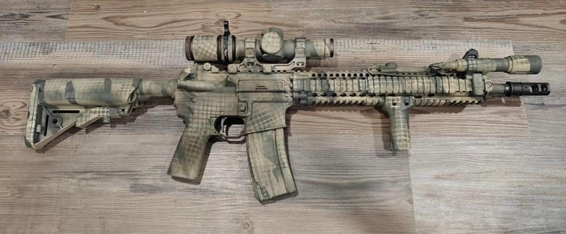 May include: A camouflaged rifle with a scope, flashlight, and vertical grip. The rifle has a magazine and is painted in shades of green, brown, and tan. The stock and handguard are also camouflaged. The rifle is lying on a wooden surface.
