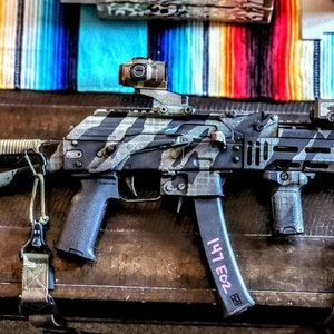 May include: A tactical firearm with a black and grey camouflage design. The weapon has a magazine with the text "147 E02" and is fitted with a sight and other accessories. A colourful textile forms the background.