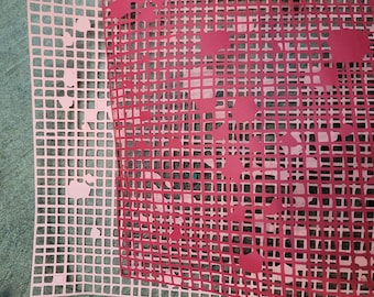 May include: Two pink and red plastic mesh sheets with a grid pattern. The sheets are layered on top of each other, with the red sheet on top. The red sheet has a pattern of cut-out shapes.