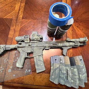 May include: A rifle with a digital camouflage pattern, alongside magazines and spray paint cans. The rifle and magazines display a green, grey, and white camouflage design. A roll of blue masking tape is also visible.