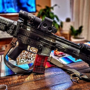 May include: A black and grey rifle with a red magazine and a scope. The rifle has a camouflage pattern and a sling. It is resting on a wooden table with a colourful striped textile.