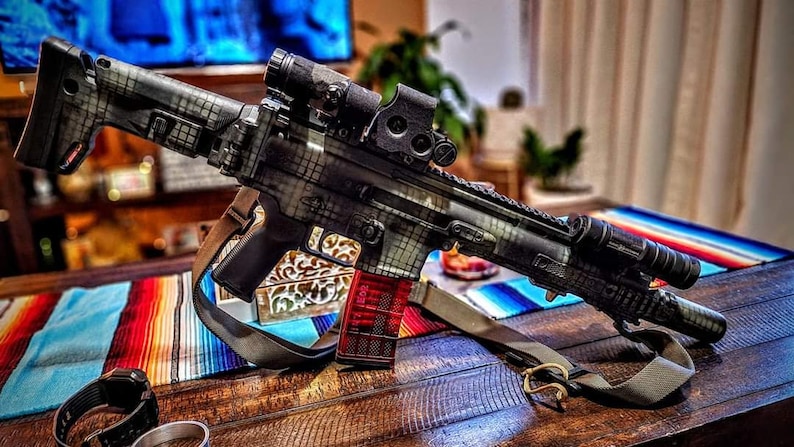 May include: A black and grey rifle with a red magazine and a scope. The rifle has a camouflage pattern and a sling. It is resting on a wooden table with a colourful striped textile.