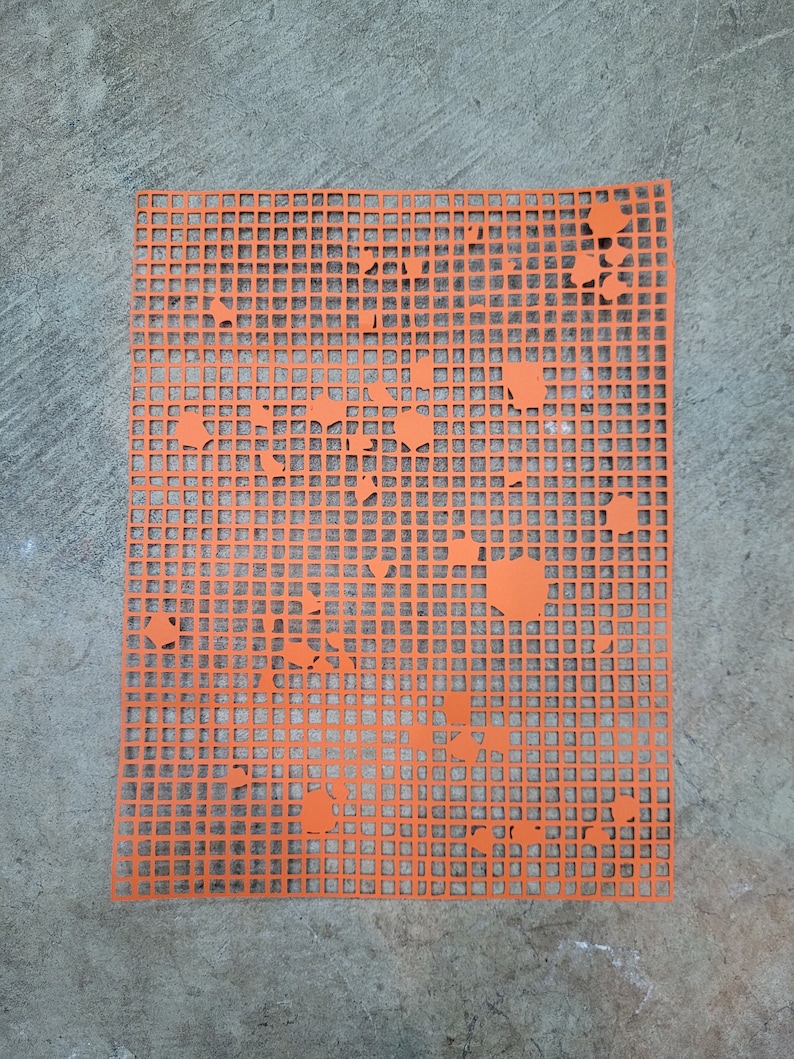 May include: A bright orange plastic canvas with a grid design and irregular cut-out shapes. The rectangular sheet is placed on a grey concrete surface. The cut-outs are of different sizes and shapes, adding a unique visual element.