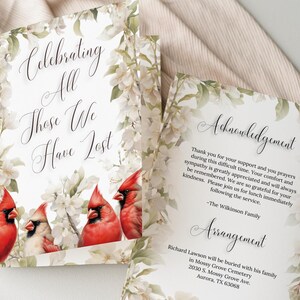 Puede incluir: Una tarjeta conmemorativa con el texto "Celebrating All Those We Have Lost" y "Acknowledgement". La tarjeta presenta un borde floral e ilustraciones de cardenales rojos. También se incluyen los detalles del arreglo.