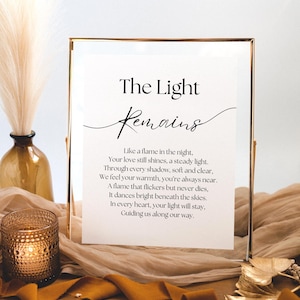 May include: A gold-framed sign with the words "The Light Remains" in elegant script. The sign features a poem below the title. A candle in a glass holder and a decorative vase are in the background, creating a warm ambiance.