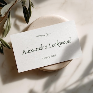 May include: A rectangular white place card featuring the name Alexandra Lockwood in a dark serif font, with a small line drawing of a lavender sprig above the name and the text TABLE ONE printed in a simple sans-serif font below the name.