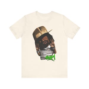 May include: A white t-shirt with a cartoon illustration of a person wearing a brown hat and a striped shirt. The person is holding their hands up in the air and the word "THUG" is written in green below them.