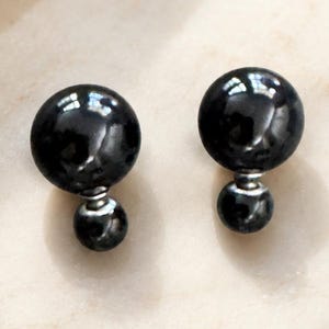May include: A pair of double-sided stud earrings featuring two glossy black spherical beads of different sizes connected by a thin silver-toned metal post. The larger bead is positioned at the front, while the smaller bead sits behind the earlobe.