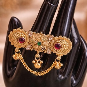 May include: A gold-toned brooch with intricate floral and circular designs, adorned with red and green gemstones. The brooch is connected by a delicate chain to dangling gold accents. The jewelry is displayed on a black hand-shaped stand.