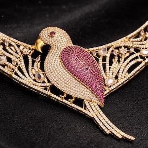 May include: A gold-toned necklace with a bird pendant. The bird has a pink breast and a gold beak, and is encrusted with small, clear gemstones. The necklace has an openwork design with clear gemstones and is set against a black background.