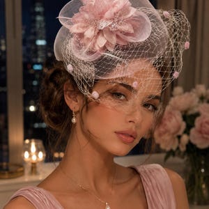 May include: A pale pink fascinator hat featuring a large fabric flower, delicate feathers, and a fine netting veil adorned with small pink pom-poms. The headpiece is worn with a gold chain necklace featuring a single teardrop pearl pendant.