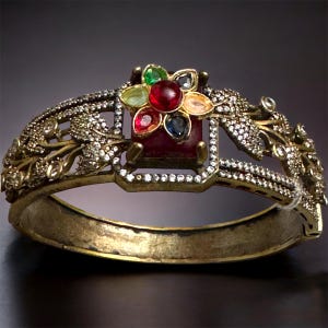 May include: Ornate gold-tone bracelet featuring a central floral design with a large red gemstone and surrounding colorful gems. The bracelet has intricate filigree and is encrusted with small, clear crystals.