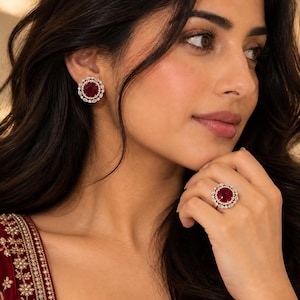 May include: A pair of round stud earrings and a matching ring, each featuring a large, deep red circular center stone surrounded by a double halo of small, clear, marquise-cut and round-cut stones set in a metallic frame with a polished finish.