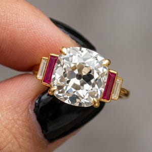 May include: A gold ring featuring a large, square-cut, clear gemstone as the centerpiece, flanked by two rectangular red gemstones and two smaller, rectangular clear gemstones. The ring is held up by a hand with black nail polish.